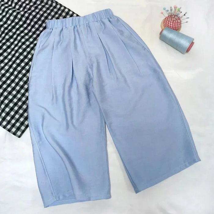 The Wide Leg Capri Pant Sewing Pattern by Longan Craft is shown on a white surface with a black-and-white checkered cloth, blue thread spool, and pincushion—download this free PDF trousers pattern for your next sewing project.