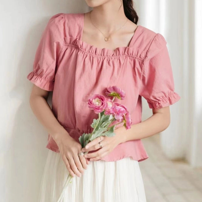 A woman wears the Longan Craft Puff Sleeve Blouse made from the free Square Neck Blouse PDF Sewing Pattern, paired with a white skirt and holding pink flowers while standing indoors against a light wall.