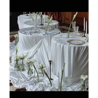 Three round tables draped with Longan Craft Soft Milk Silk Fabric in caramel, accented by white roses in vases, glassware, plates, and tall candles for a minimalist wedding setting.