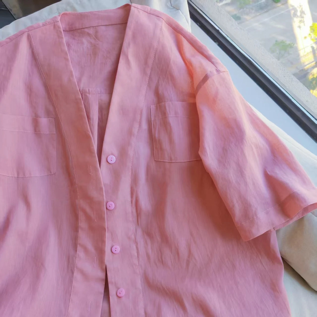 A Women's V-neck Oversize Shirt, made using the Longan Craft PDF sewing pattern, is laid flat near a sunlit window. The light pink, short-sleeved button-up has two chest pockets.
