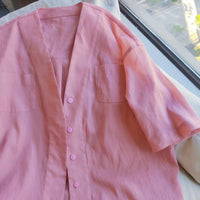 A Women's V-neck Oversize Shirt, made using the Longan Craft PDF sewing pattern, is laid flat near a sunlit window. The light pink, short-sleeved button-up has two chest pockets.