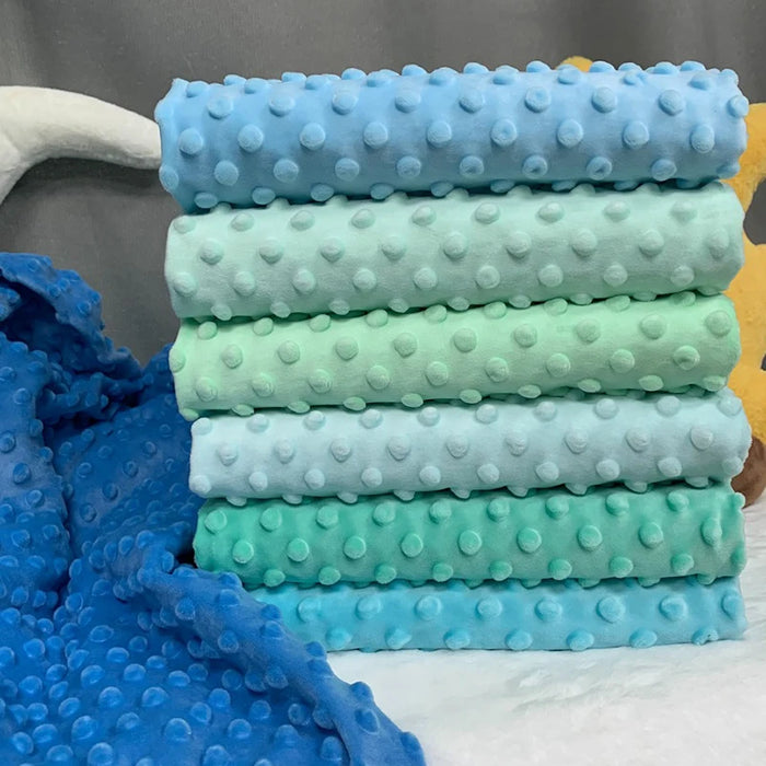 A stack of six pastel blankets crafted from Longon Craft's Dotted Minky Fabric By The Yard in shades of blue, green, and turquoise. A plush blue blanket made with this soft 3D dot minky material is draped to the side.