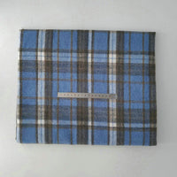 A piece of Multicolor Plaid Mammoth Flannel Fabric by Longan Craft, featuring white, black, and brown stripes on blue, is folded under a metal ruler laid horizontally across its center on a light surface.