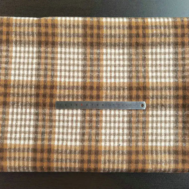 A metal ruler rests on Longan Craft’s Multicolor Plaid Mammoth Flannel Fabric, showcasing the brown and cream checkered polyester cotton blend—ideal for comfy shirts.