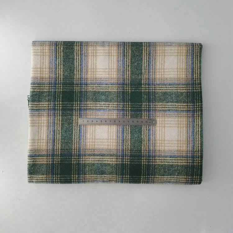 A folded Longan Craft Multicolor Plaid Mammoth Flannel Fabric by the yard rests on a white surface with a metal ruler for scale, showing its green, beige, and blue brushed polyester cotton textile, perfect for comfy shirts.