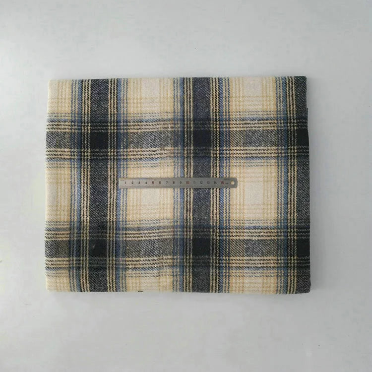 A folded Multicolor Plaid Mammoth Flannel Fabric by Longan Craft in beige, black, and blue rests on a white surface with a metal ruler laid horizontally across the center for scale.