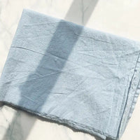 A piece of light blue 100% cotton crepe fabric from Longan Craft, known for its pure color and breathable washed crepe material ideal for casual wear and clothing, lies flat on a white marble surface. Sunlight casts a rectangular shadow across the slightly rough-textured fabric, which features a frayed bottom edge.