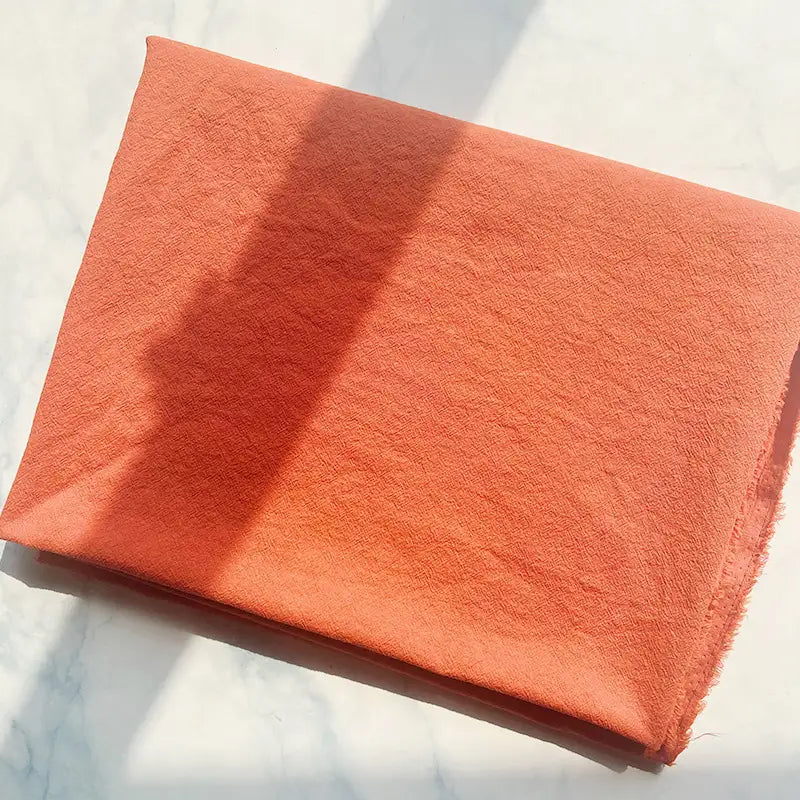 A piece of Longan Craft 100% Cotton Crepe Fabric, in a pure orange color, is folded neatly on a light surface, with gentle natural light and soft shadows highlighting its breathable texture.