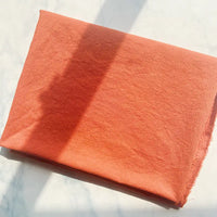 A piece of Longan Craft 100% Cotton Crepe Fabric, in a pure orange color, is folded neatly on a light surface, with gentle natural light and soft shadows highlighting its breathable texture.