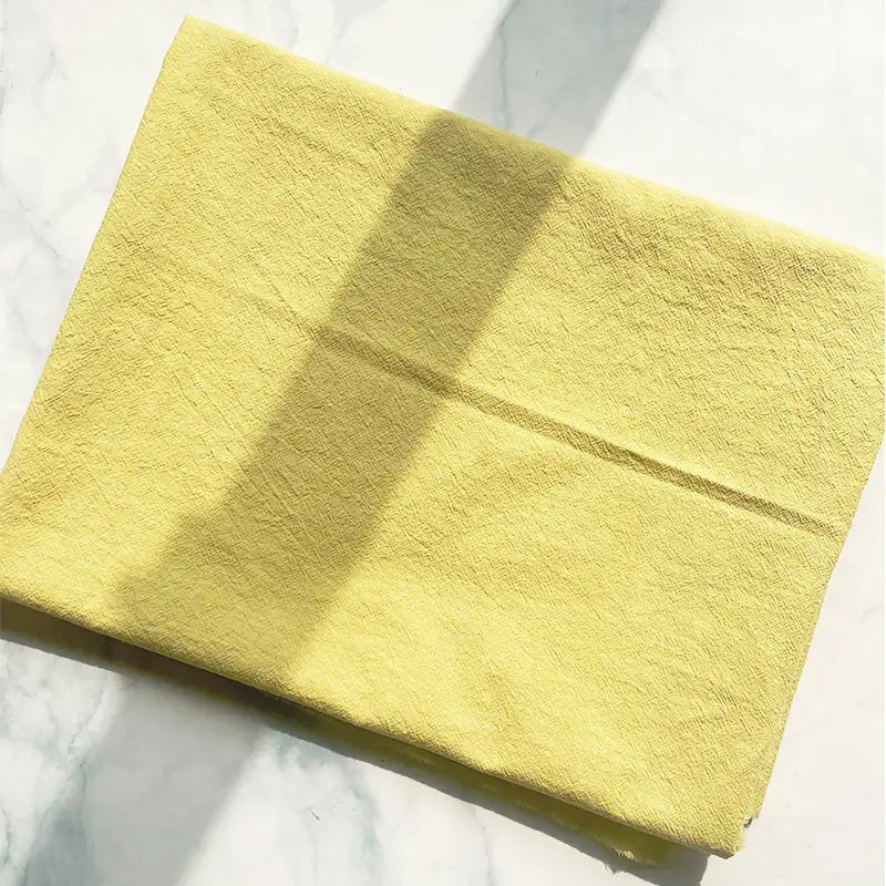 A folded piece of Longan Craft 100% Cotton Crepe Fabric, in a pure yellow shade, rests on a white marble surface with sunlight casting gentle shadows across it.