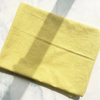 A folded piece of Longan Craft 100% Cotton Crepe Fabric, in a pure yellow shade, rests on a white marble surface with sunlight casting gentle shadows across it.