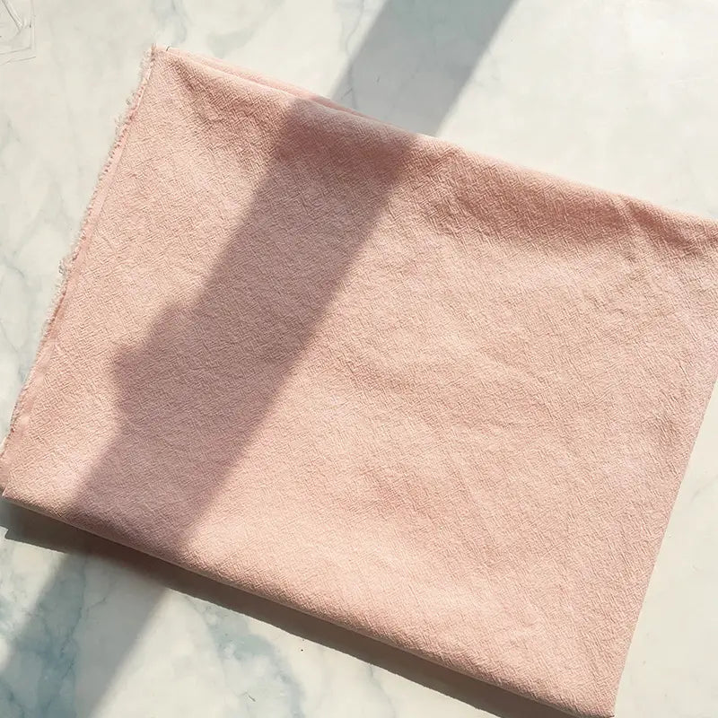 A neatly folded length of Longan Craft 100% Cotton Crepe Fabric By The Yard, pure color washed crepe material for clothing, rests on a white surface with sunlight casting a soft shadow. The breathable fabric’s textured finish is visible, and the left edge appears slightly frayed.