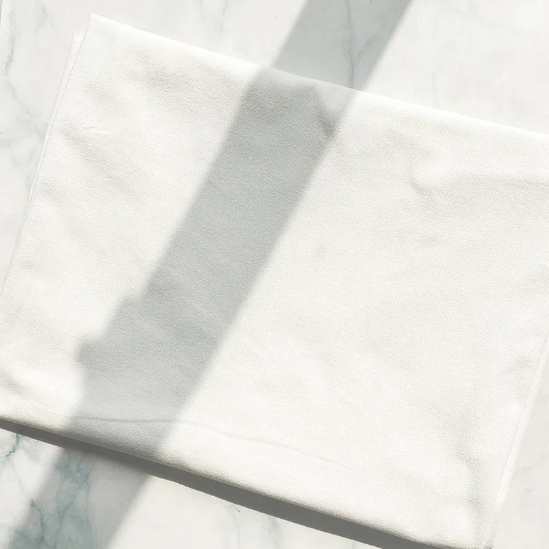 A piece of white 100% cotton crepe fabric from Longan Craft, known for its pure color and breathable texture, lies folded on a light marble surface, partially veiled in shadow.