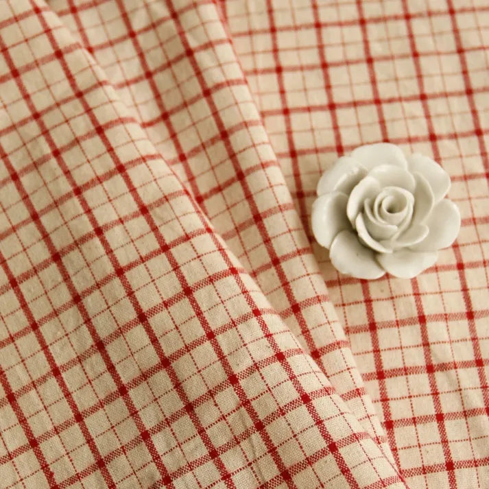 A piece of Longan Craft’s 100% Cotton Gingham Poplin Fabric, featuring a multicolor yarn-dyed checkered pattern, is elegantly draped with a small white ceramic rose resting on top.