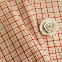 A piece of Longan Craft’s 100% Cotton Gingham Poplin Fabric, featuring a multicolor yarn-dyed checkered pattern, is elegantly draped with a small white ceramic rose resting on top.