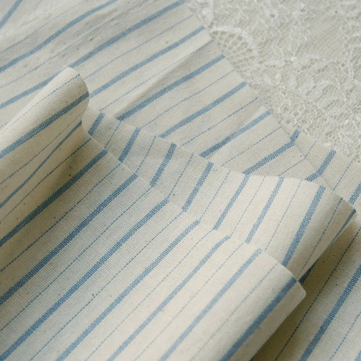 A close-up view of the Longan Craft 100% Cotton Gingham Poplin Fabric By The Yard showcases its light beige background with blue vertical stripes and multicolor yarn-dyed checkered pattern, neatly folded on a white lace surface. The image emphasizes the premium soft texture and distinctive gingham design of this poplin material, ideal for garment making.