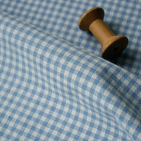 A wooden spool rests on a slightly wrinkled piece of Longan Craft 100% Cotton Gingham Poplin Fabric, featuring a light blue and white checkered pattern, spread out beneath it.
