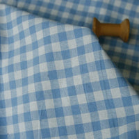 The 100% Cotton Gingham Poplin Fabric by the yard from Longan Craft, featuring a multicolor yarn-dyed checkered pattern in light blue and white, is spread out with a small wooden spool partially visible near the top right corner.