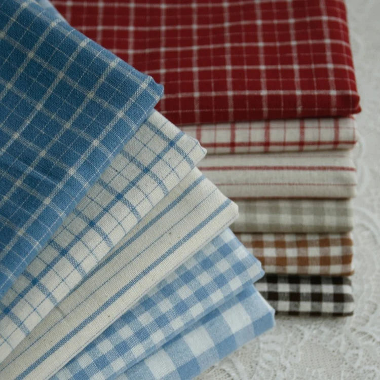 A neat stack of Longan Craft 100% Cotton Gingham Poplin Fabric By The Yard, featuring multicolor yarn-dyed checkered patterns in shades of blue, red, brown, gray, and black, is arranged on a white lace surface—perfect premium soft poplin material for garment making.