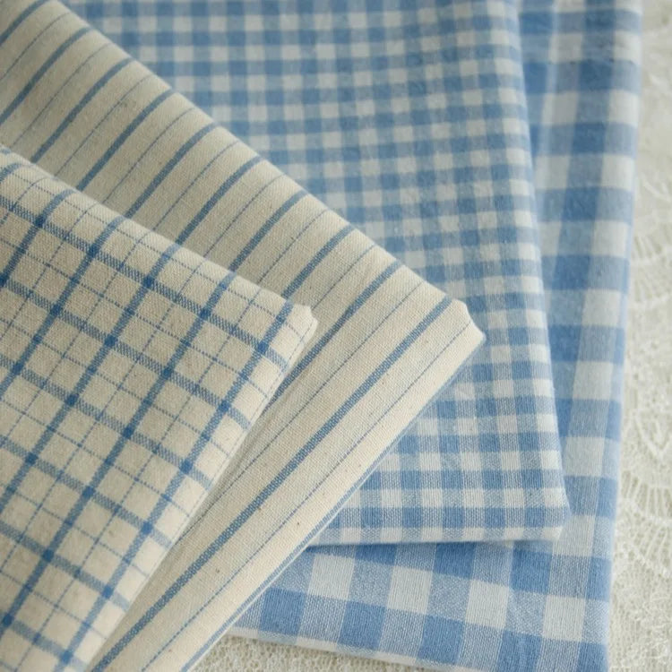 Four folded pieces of Longan Craft 100% Cotton Gingham Poplin Fabric, featuring various blue and white checkered designs, are laid on a white lace surface.