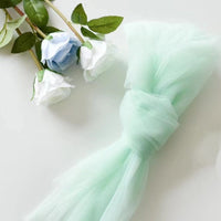 Two white and one light blue artificial rose rest beside a bundle of mint green Fine Mesh Material By The Yard nylon mesh fabric from Longan Craft, displayed on a white surface.
