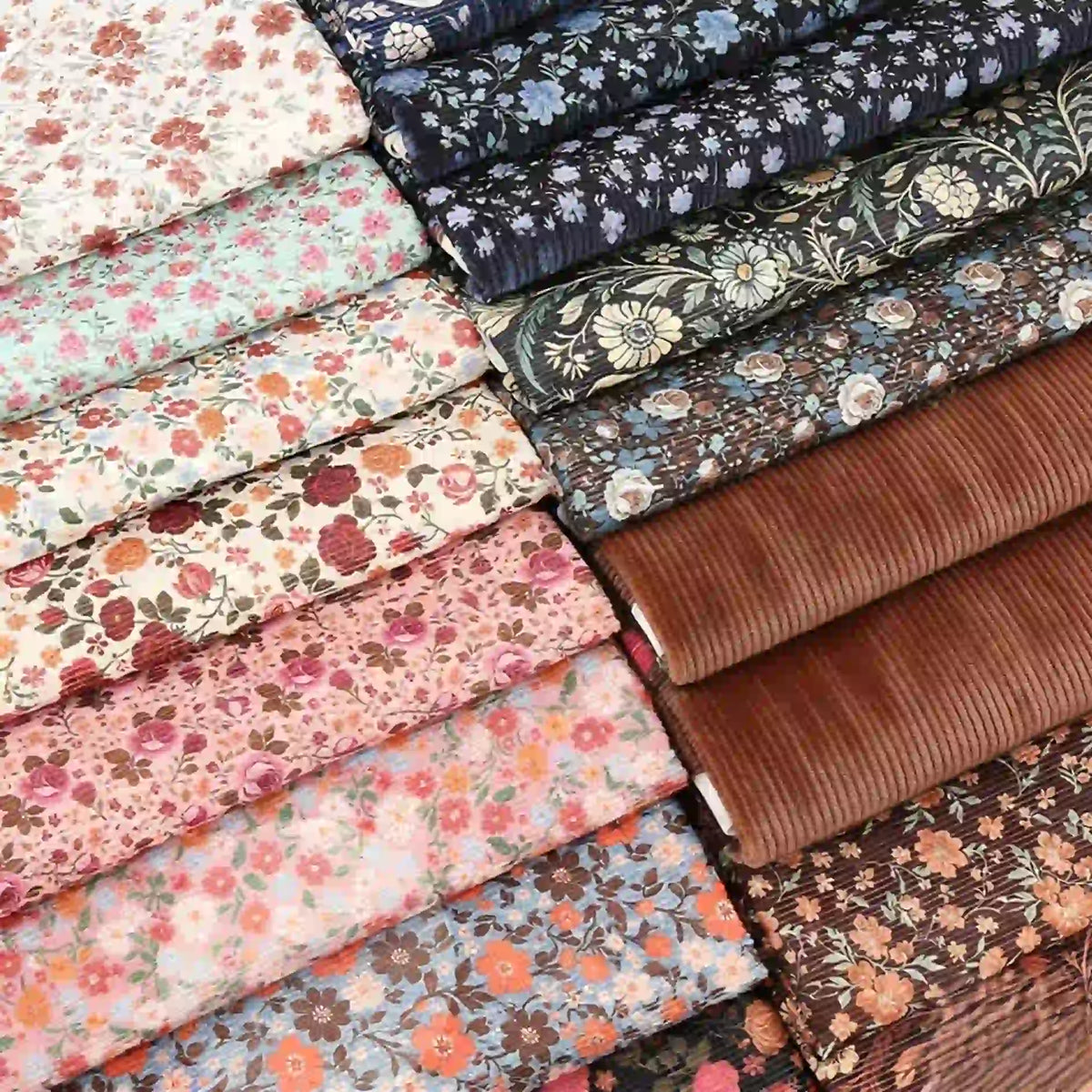Neatly arranged stacks of Longan Craft 16 Wale Floral Printed Corduroy Fabric by the yard show colorful fine wale corduroy in floral patterns and shades of brown, blue, pink, and cream—perfect for casual wear and premium textile projects.