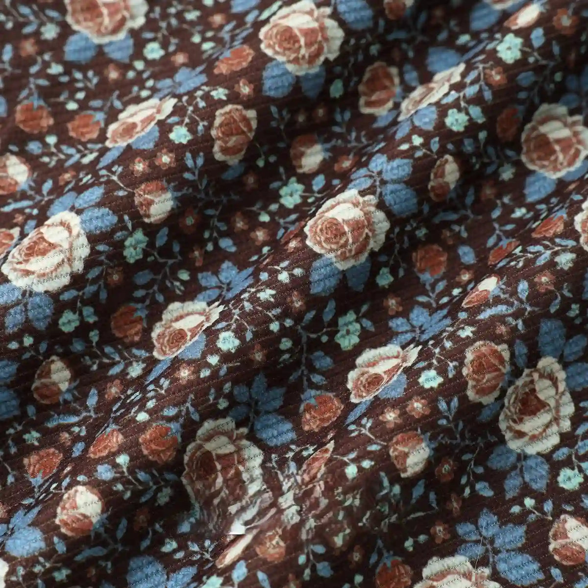 Close-up of Longan Craft 16 Wale Floral Printed Corduroy Fabric By The Yard, showing cream, blue, and rusty red roses with green leaves. Gentle folds add soft shadows and texture to this colorful fine wale premium corduroy textile.