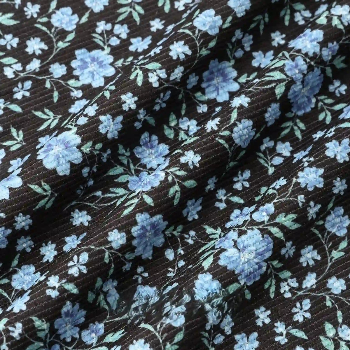 A close-up of Longan Craft's 16 Wale Floral Printed Corduroy Fabric shows fine black corduroy with small blue and light blue flowers, green leaves, and soft folds—premium material by the yard for casual wear.
