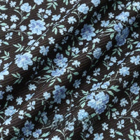 A close-up of Longan Craft's 16 Wale Floral Printed Corduroy Fabric shows fine black corduroy with small blue and light blue flowers, green leaves, and soft folds—premium material by the yard for casual wear.