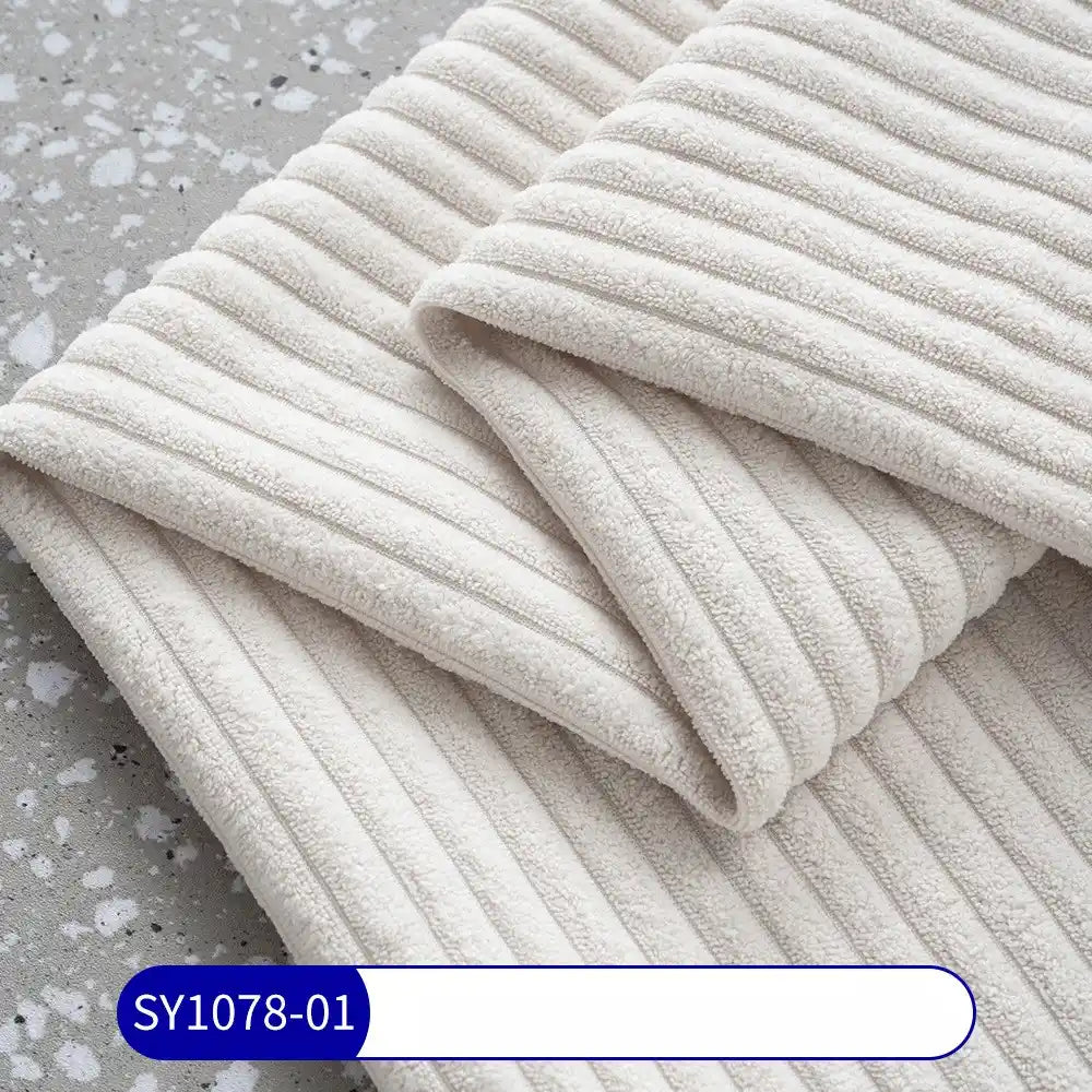 A folded piece of Longan Craft’s 2.5 Wale Corduroy Fabric By The Yard in cream lies on a gray terrazzo surface, showing off its soft, ribbed texture. A blue and white label at the bottom reads SY1078-01.
