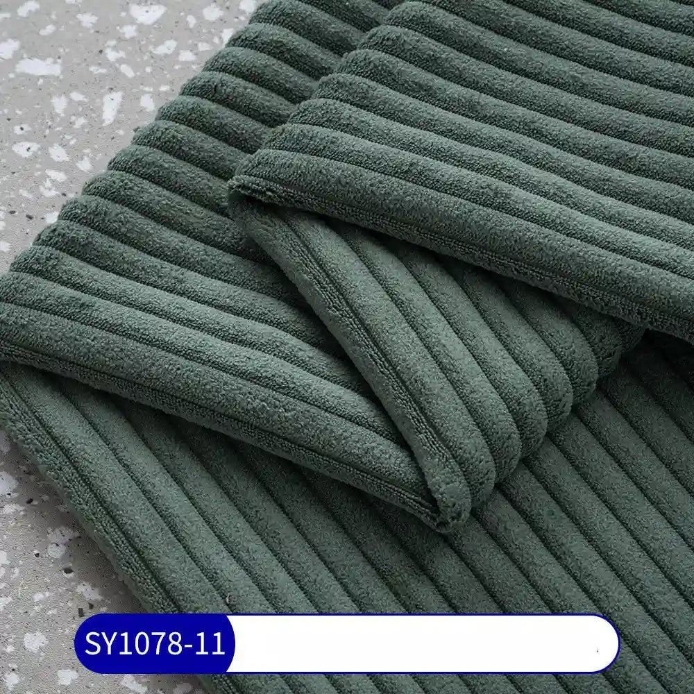A close-up of folded dark green towels, made from Longan Craft's 2.5 Wale Corduroy Fabric By The Yard (Multicolor Polyester Corduroy Upholstery Material), resting on a gray speckled surface. A blue and white label shows SY1078-11.