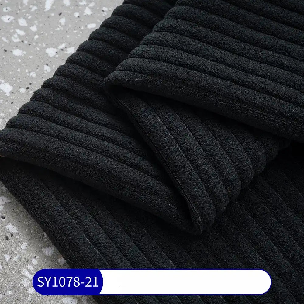 A close-up of folded, ribbed black 2.5 Wale Corduroy Fabric By The Yard from Longan Craft on a gray speckled surface. A blue and white label at the bottom shows SY1078-21.