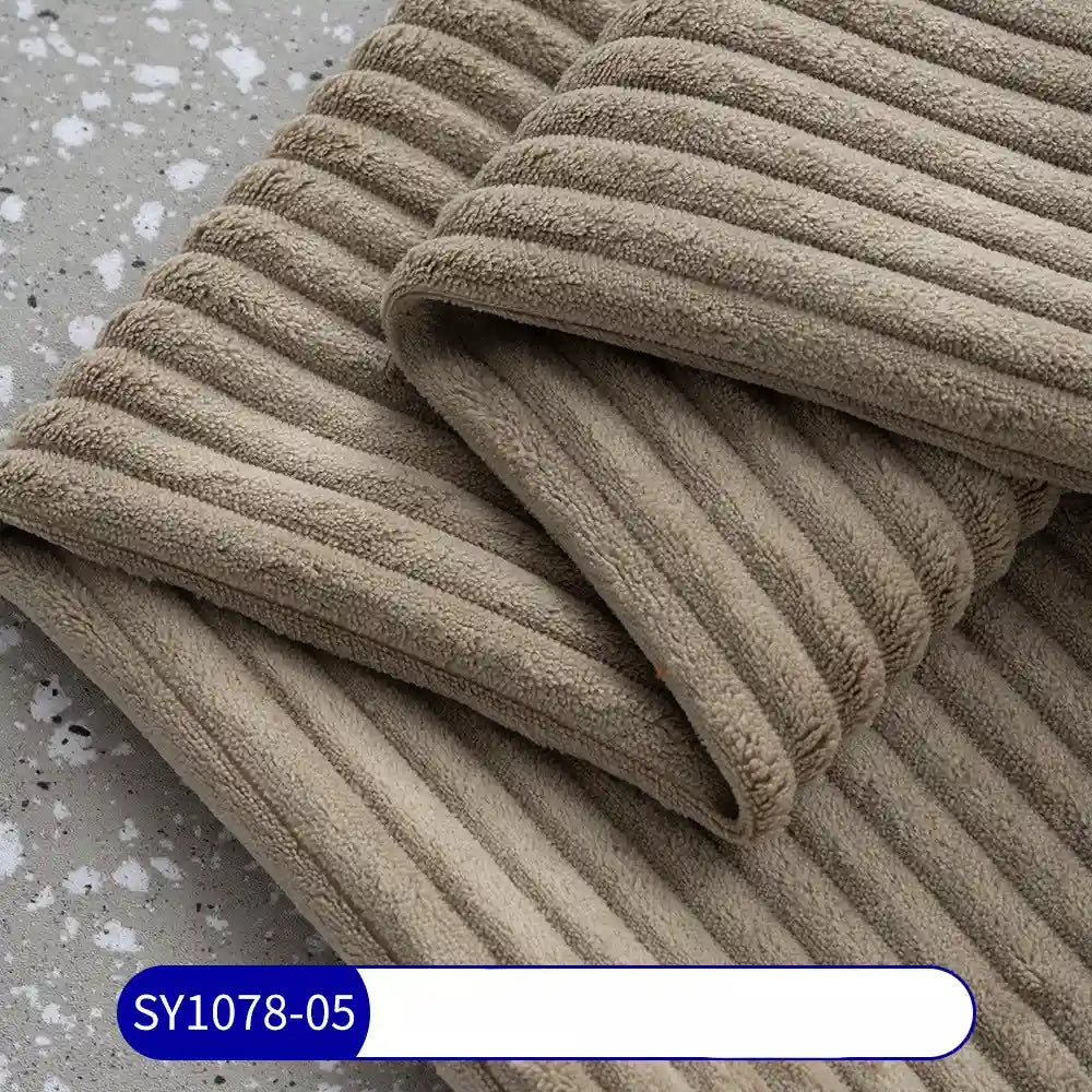 A close-up of folded, ribbed beige towels on a speckled gray surface highlights the rich texture of Longan Craft’s 2.5 Wale Corduroy Fabric By The Yard, Multicolor Polyester Upholstery Material. A label at the bottom reads SY1078-05 on blue and white.