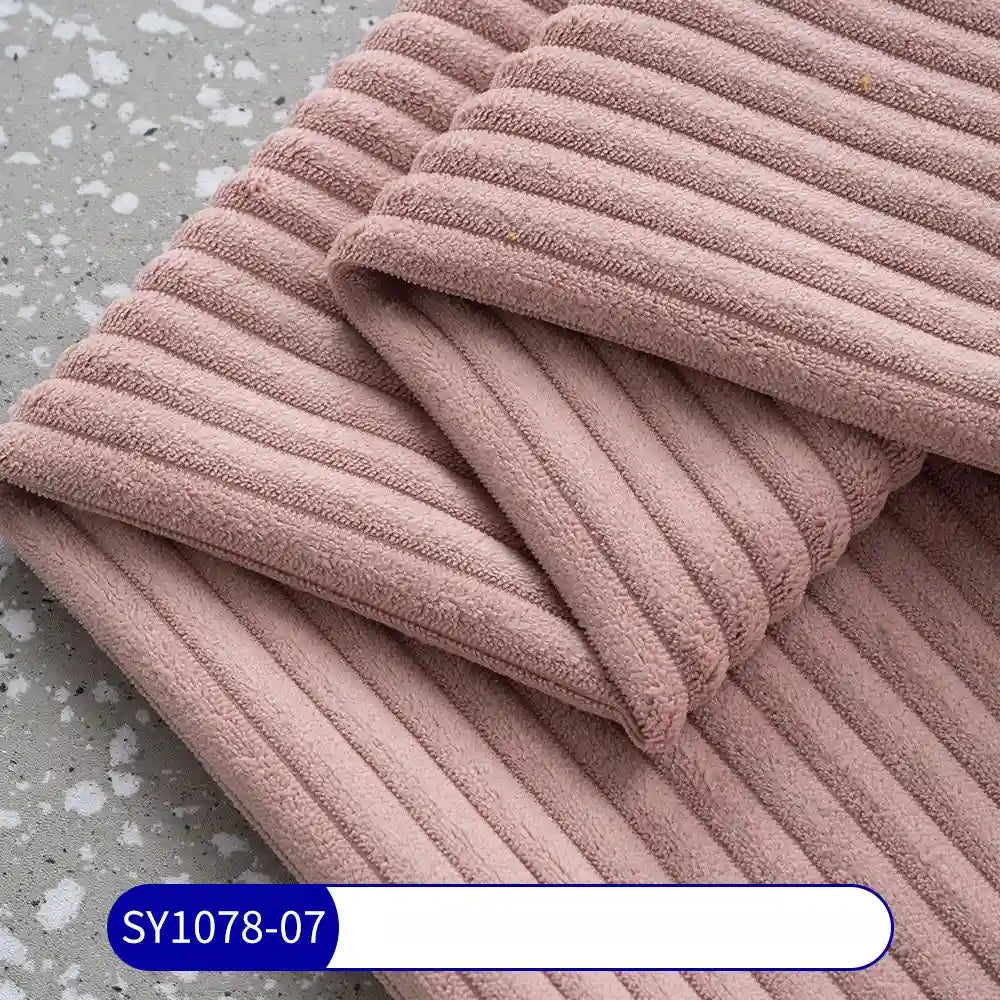 A close-up of folded, ribbed light mauve 2.5 Wale Corduroy Fabric By The Yard from Longan Craft on a gray speckled surface, with a blue SY1078-07 label. Made of multicolor polyester corduroy material for upholstery.