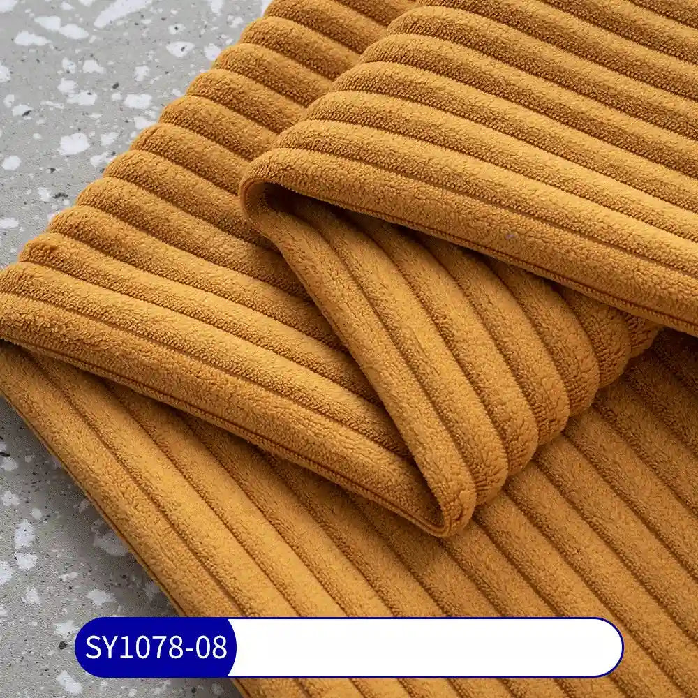 A close-up of mustard-yellow, ribbed Longan Craft 2.5 Wale Corduroy Fabric by the yard shows its soft texture on a gray surface. A blue and white SY1078-08 label highlights this multicolor polyester corduroy, ideal for upholstery projects.