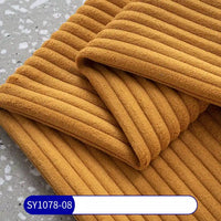 A close-up of mustard-yellow, ribbed Longan Craft 2.5 Wale Corduroy Fabric by the yard shows its soft texture on a gray surface. A blue and white SY1078-08 label highlights this multicolor polyester corduroy, ideal for upholstery projects.