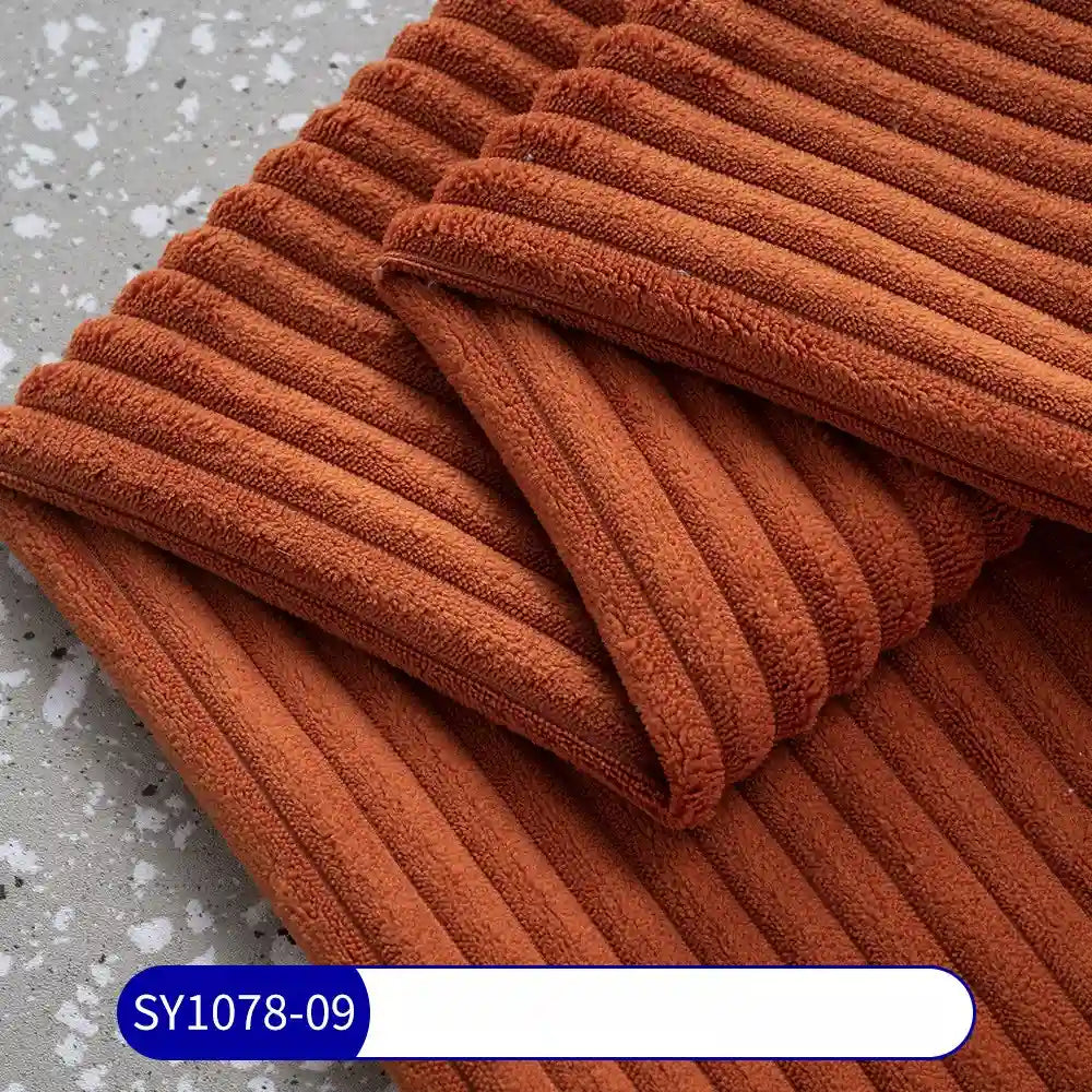 Close-up of a folded, ribbed burnt orange Longan Craft 2.5 Wale Corduroy Fabric By The Yard on a speckled gray surface. A label at the bottom reads “SY1078-09” in white text on a blue background.
