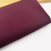 A close-up of Longan Craft’s Solid Color 21 Wales Corduroy Fabric By The Yard in maroon, fine wale cotton material, folded on a white surface with a wooden edge in the background.