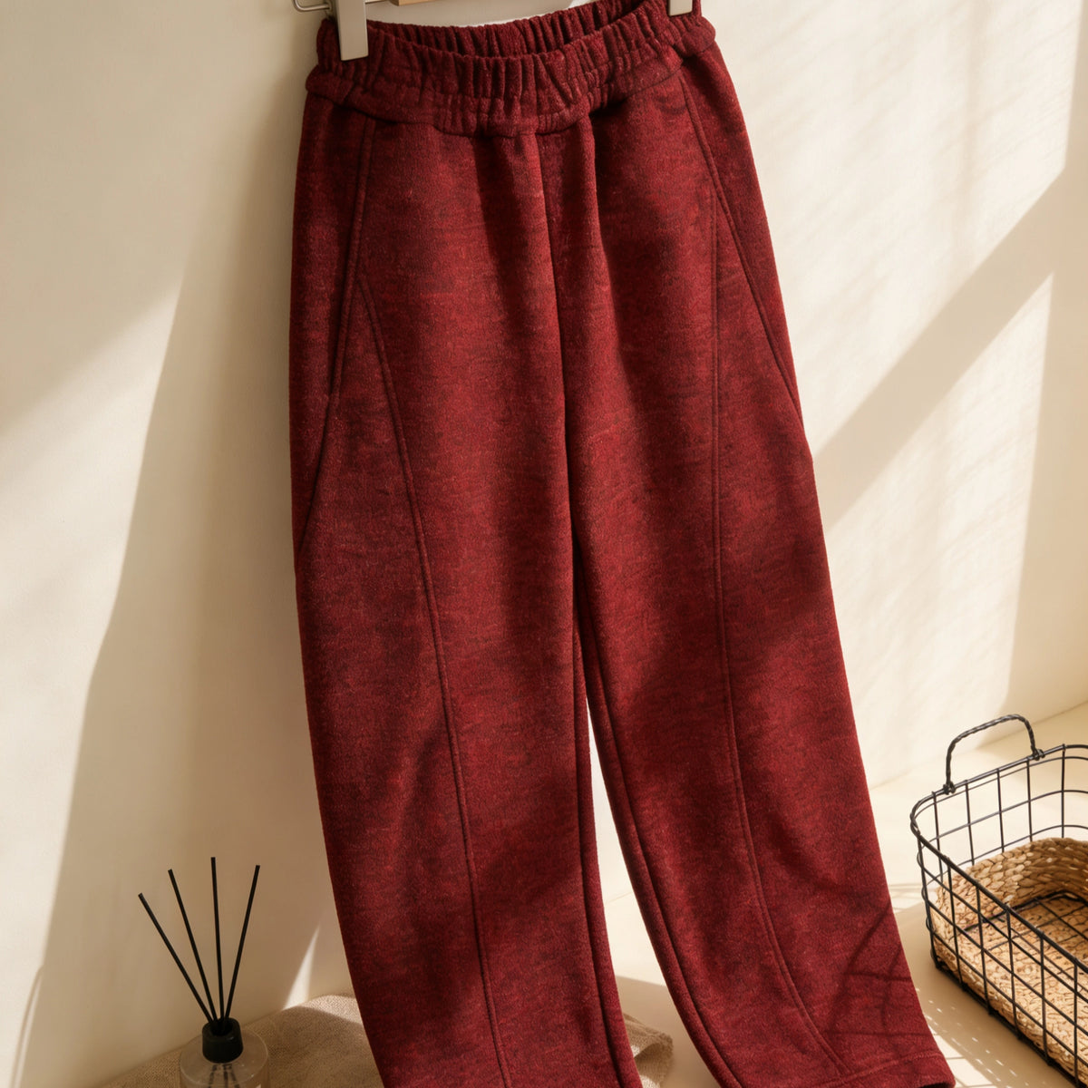 A pair of red high-waisted tailored pants made using the Structured Pants Sewing Pattern by Longan Craft hangs on a hanger in a sunlit room, beside a wire basket and reed diffuser. Free PDF pattern download available.