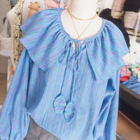 A mannequin showcases the Ruffle Collar Long-Sleeved Blouse made with Longan Craft’s free PDF sewing pattern, featuring colorful stripes and a tasseled tie—perfect inspiration for your next sewing project. Fabric rolls complete the stylish scene.