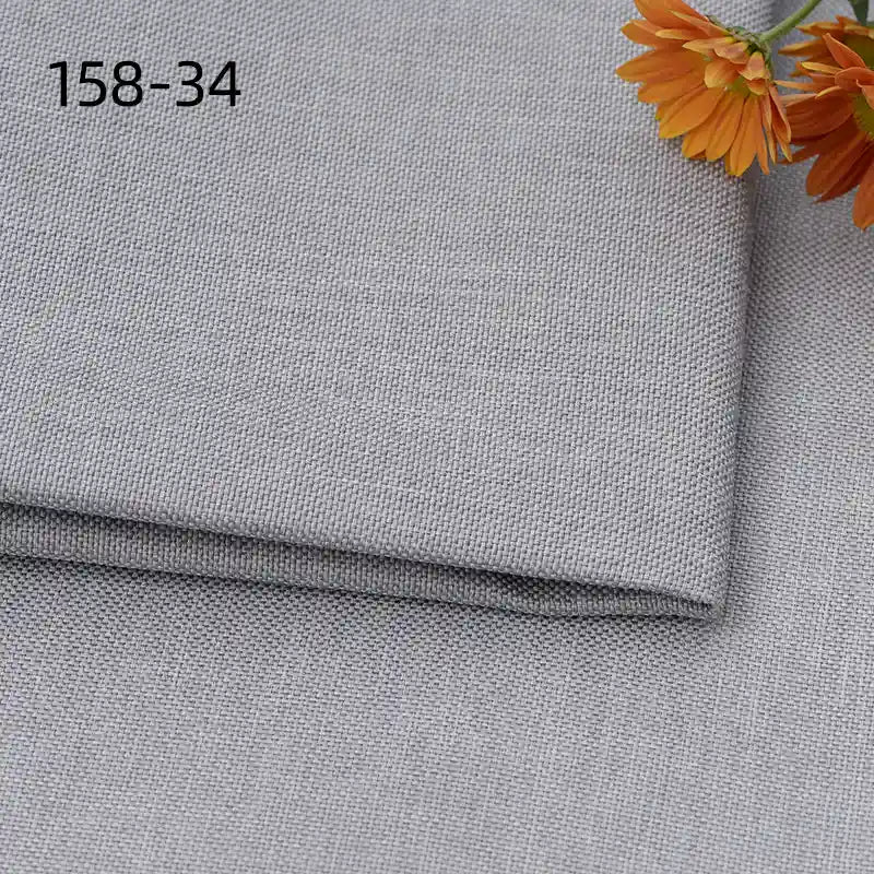 A close-up of Longan Craft’s faux linen upholstery fabric by the yard shows folded light gray polyester with woven texture. Orange flowers appear in the top right, and “158-34” is printed in black on the top left of the fabric.