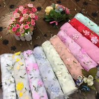 Several rolls of Longan Craft 3D Floral Embroidered Tulle Fabric By The Yard—colorful tulle material with botanical embroidery for bridal use—are displayed on a table, alongside artificial flower bouquets and decorative accents.