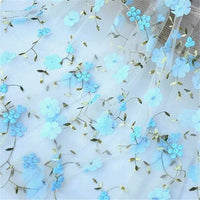 Longan Craft’s 3D Floral Embroidered Tulle Fabric By The Yard features sheer white tulle adorned with blue flowers and greenish-gold stems—ideal botanical embroidery for airy, elegant wedding dress designs.