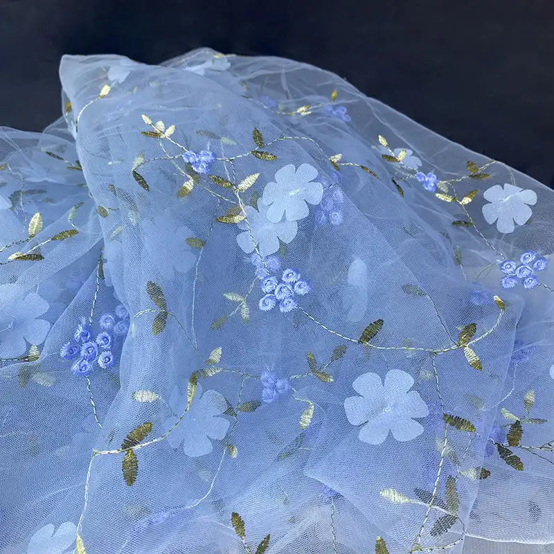 Longan Craft’s 3D Floral Embroidered Tulle Fabric by the yard features colorful botanical embroidery with white flowers, blue blooms, and gold leaves—ideal for bridal gowns, as shown against a dark background.