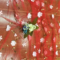 A piece of Longan Craft 3D Floral Embroidered Tulle Fabric By The Yard, featuring colorful botanical embroidery, is displayed on wood. At the center, a small bouquet of artificial flowers with green leaves and cream and blue blossoms adds charm.