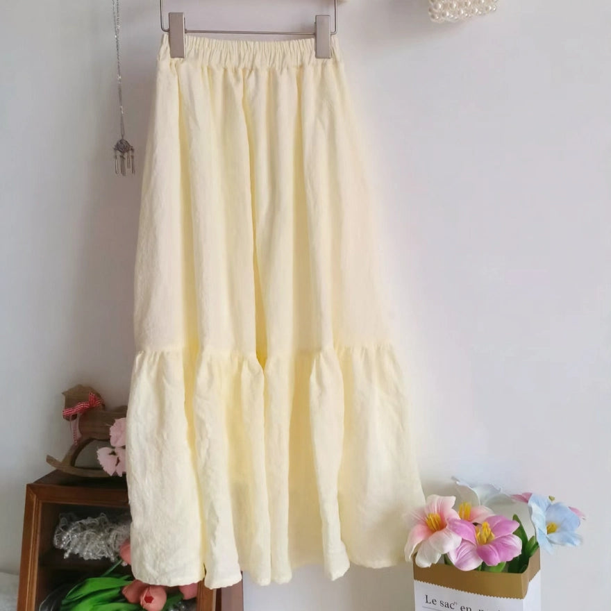 A long, light yellow skirt hangs on a hanger by a shelf with pink flowers and a box of pastel blooms—perfect inspiration for your next handmade project using the Women’s Pleated Midi Skirt Sewing Pattern by Longan Craft. Free PDF download available.