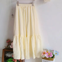 A long, light yellow skirt hangs on a hanger by a shelf with pink flowers and a box of pastel blooms—perfect inspiration for your next handmade project using the Women’s Pleated Midi Skirt Sewing Pattern by Longan Craft. Free PDF download available.