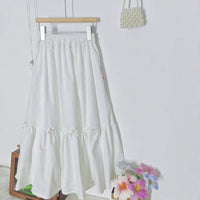 Showcasing the Women’s Pleated Midi Skirt Sewing Pattern by Longan Craft—a free PDF for your handmade wardrobe—displayed on a wooden hanger with a pearl handbag and bright faux flowers nearby against a clean white wall.