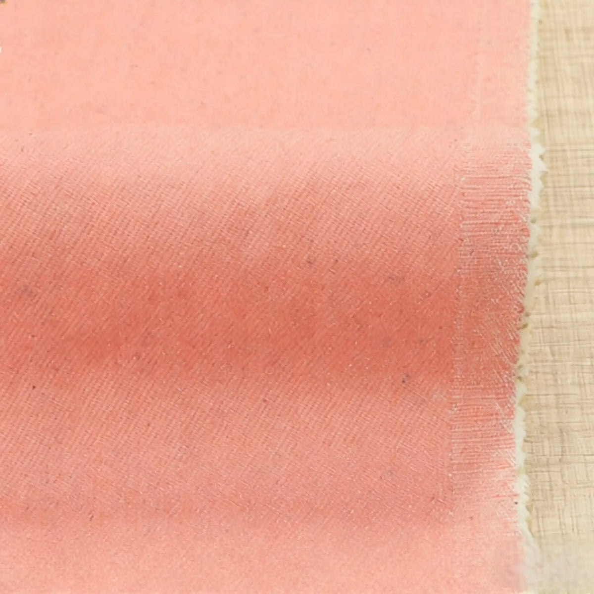 A close-up of Longan Craft's 9.5oz Stretch Denim Fabric in light pink, featuring a textured diagonal weave and slightly frayed edges, displayed on a wooden surface—an ideal solid cotton material for jeans, sold by the yard.