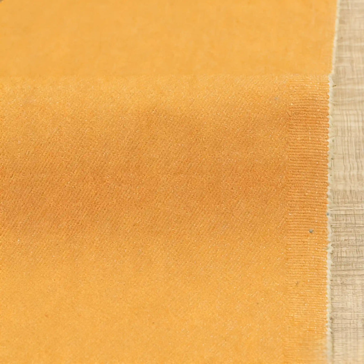 A close-up of Longan Craft's 9.5oz Stretch Denim Fabric in orange, a medium-weight cotton denim with a soft, velvety texture and slightly frayed edges, laid flat on a beige woven surface—ideal for making jeans.